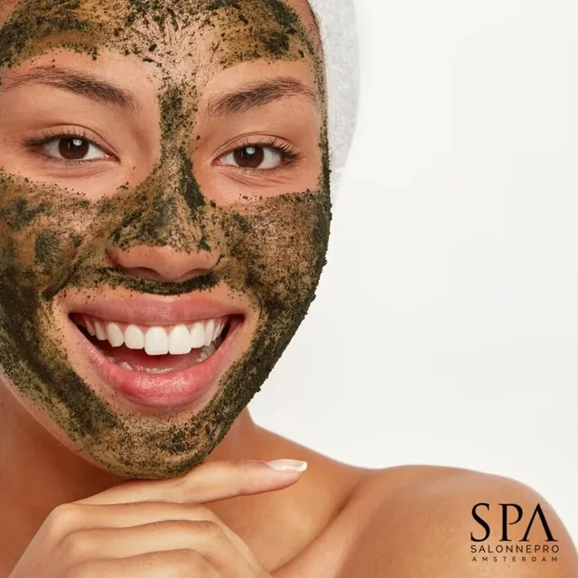 Een lachende vrouw met een groen kruidenmasker op het gezicht.